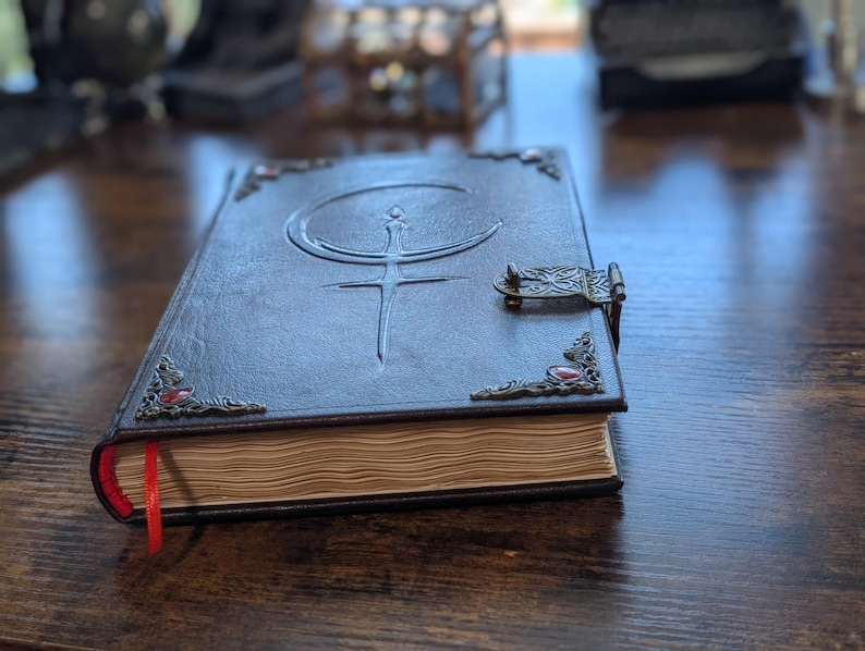 Puede incluir: Un diario de cuero marr&oacute;n oscuro con un cierre decorativo y adornos en las esquinas. La cubierta presenta un dise&ntilde;o en relieve de una luna creciente y una espada. Una cinta roja de marcador es visible, y las p&aacute;ginas tienen un aspecto envejecido.