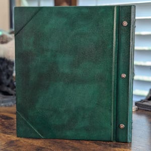 May include: A dark green leather-bound book with a textured surface. The book has a spine with metal rivets and a diagonal line detail on the cover. The book is sitting on a wooden surface.