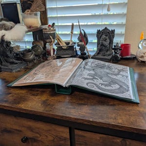 May include: An open book with a green cover and detailed illustrations of a dragon and handwritten text. The book is on a dark wooden desk, surrounded by various decorative items, including candles and figurines, creating a mystical atmosphere.