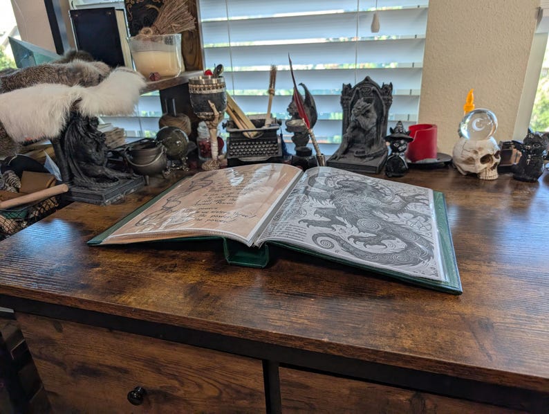 May include: An open book with a green cover and detailed illustrations of a dragon and handwritten text. The book is on a dark wooden desk, surrounded by various decorative items, including candles and figurines, creating a mystical atmosphere.