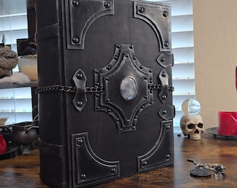 May include: A large, black, gothic-style book with metal detailing. The book has decorative metal corner pieces, a central oval embellishment, and a chain closure. The cover has a textured appearance, possibly leather or a similar material.