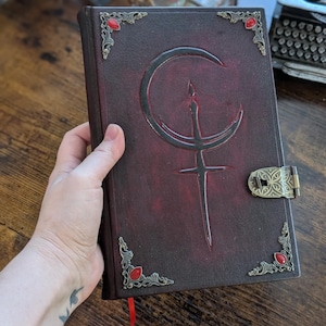 Puede incluir: Un diario de cuero burdeos oscuro con un cierre y esquinas decorativas de lat&oacute;n. La cubierta presenta un dise&ntilde;o en relieve con una media luna y una espada. Piedras preciosas rojas est&aacute;n engastadas en las esquinas, y una cinta roja de marcador es visible.