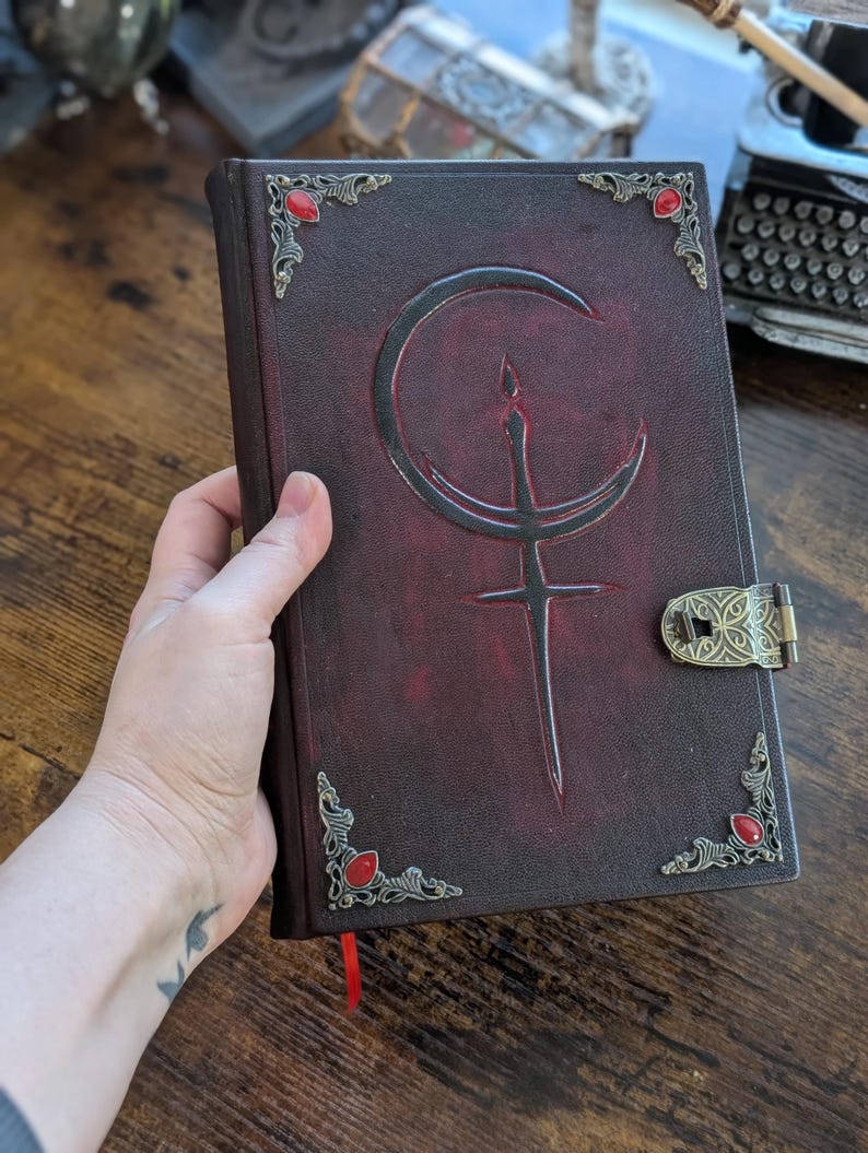 Puede incluir: Un diario de cuero burdeos oscuro con un cierre y esquinas decorativas de lat&oacute;n. La cubierta presenta un dise&ntilde;o en relieve con una media luna y una espada. Piedras preciosas rojas est&aacute;n engastadas en las esquinas, y una cinta roja de marcador es visible.