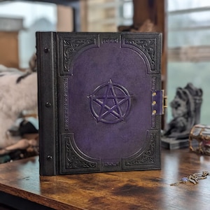 May include: A dark purple and black leather-bound book with a pentagram embossed on the cover. The book has ornate corner designs and a clasp closure. The book is sitting on a wooden surface.
