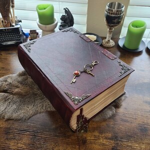 K&ouml;nnte beinhalten: Ein gro&szlig;es, burgunderfarbenes, ledergebundenes Buch mit verzierten Metallapplikationen und einem Schl&uuml;sselanh&auml;nger an der Umschlagseite. Das Buch ist geschlossen und liegt auf einer Holzoberfl&auml;che.