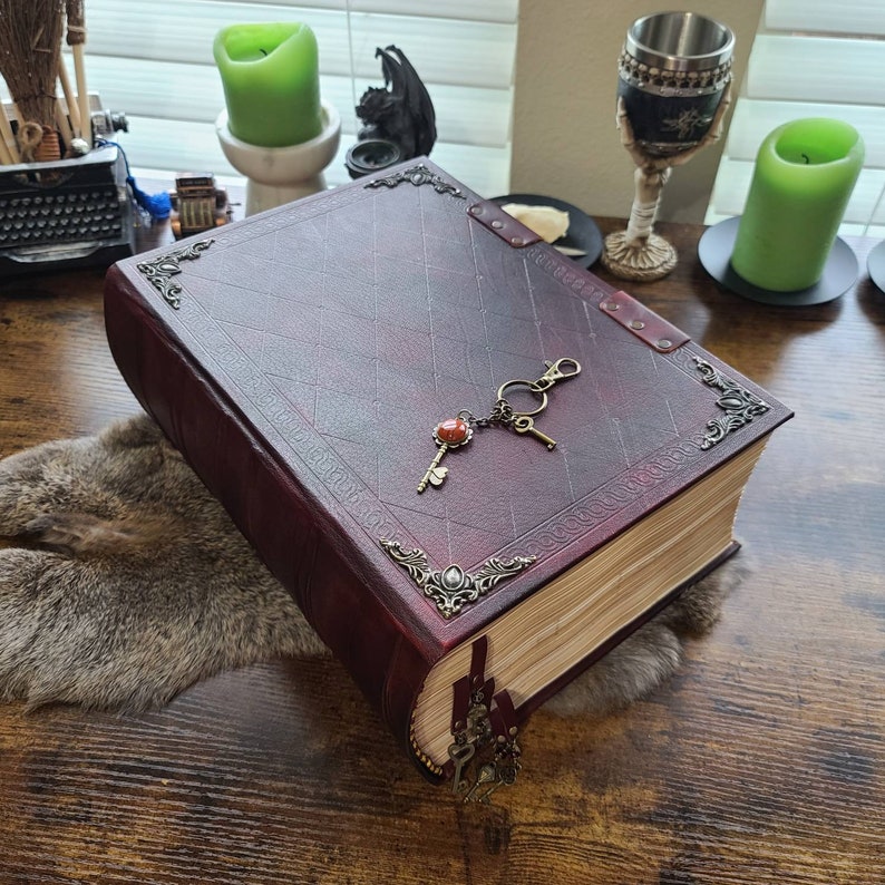 K&ouml;nnte beinhalten: Ein gro&szlig;es, burgunderfarbenes, ledergebundenes Buch mit verzierten Metallapplikationen und einem Schl&uuml;sselanh&auml;nger an der Umschlagseite. Das Buch ist geschlossen und liegt auf einer Holzoberfl&auml;che.