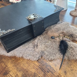 May include: A large, black, leather-bound book with decorative metal corners and a clasp. A key chain with a key and a black feather lies on a faux fur surface, evoking a gothic or fantasy aesthetic.