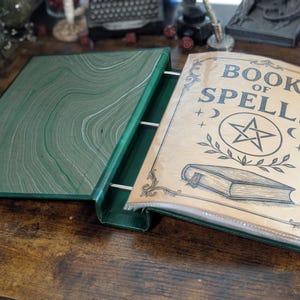 May include: Open green binder with the words "BOOK OF SPELLS" printed on the cover. The cover features a pentagram, crescent moons, and a book illustration. The binder is open, revealing a green and white marbled interior.