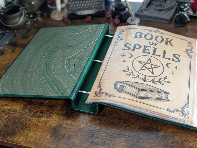 May include: Open green binder with the words "BOOK OF SPELLS" printed on the cover. The cover features a pentagram, crescent moons, and a book illustration. The binder is open, revealing a green and white marbled interior.