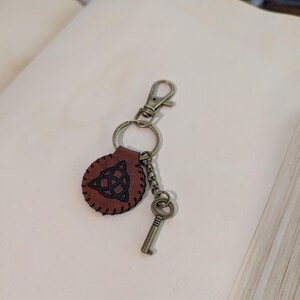 May include: A brown leather keychain with a Celtic knot design, attached to a metal clasp and key. The keychain is round with a stitched border. The key and clasp are a bronze color.