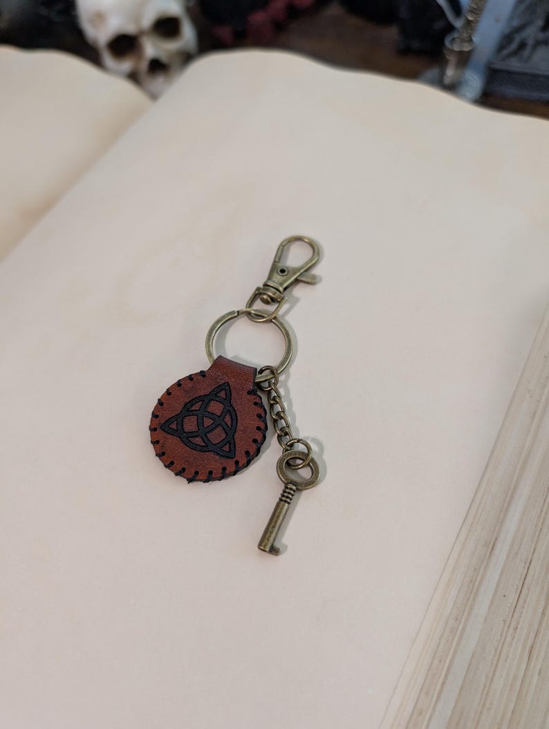 May include: A brown leather keychain with a Celtic knot design, attached to a metal clasp and key. The keychain is round with a stitched border. The key and clasp are a bronze color.