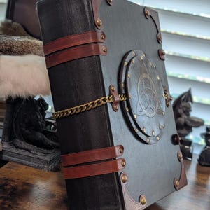 May include: A large, black leather-bound journal with brown leather straps and corner accents. A brass chain secures the journal, and a circular emblem with a Celtic knot design adorns the cover. The journal has a vintage, gothic aesthetic.