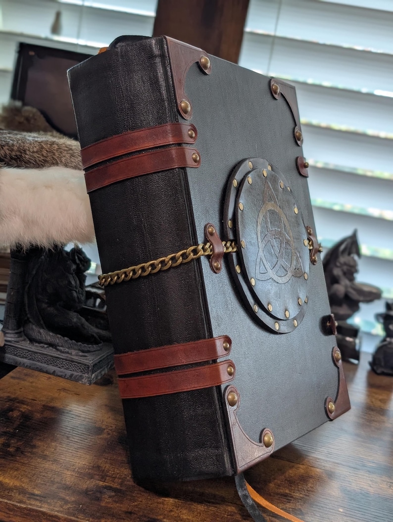 May include: A large, black leather-bound journal with brown leather straps and corner accents. A brass chain secures the journal, and a circular emblem with a Celtic knot design adorns the cover. The journal has a vintage, gothic aesthetic.