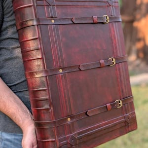 Peut inclure: Grand livre relié en cuir bordeaux avec trois sangles en cuir marron et boucles en laiton. Le livre présente un motif en losange décoratif sur les coins et des lignes verticales. Le livre est tenu par une personne.
