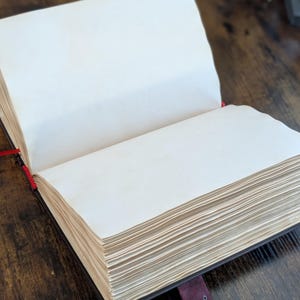 Puede incluir: Un diario abierto con p&aacute;ginas gruesas de color crema. El diario tiene una cubierta oscura con una correa burdeos y un cierre plateado. Una cinta roja como marcador es visible. El diario est&aacute; sobre una superficie de madera oscura.