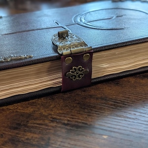 Puede incluir: Un diario de cuero marr&oacute;n oscuro con un cierre y adornos de esquina de metal decorativos. La cubierta presenta un dise&ntilde;o en relieve y las p&aacute;ginas est&aacute;n envejecidas. El diario est&aacute; cerrado con una correa de cuero burdeos.