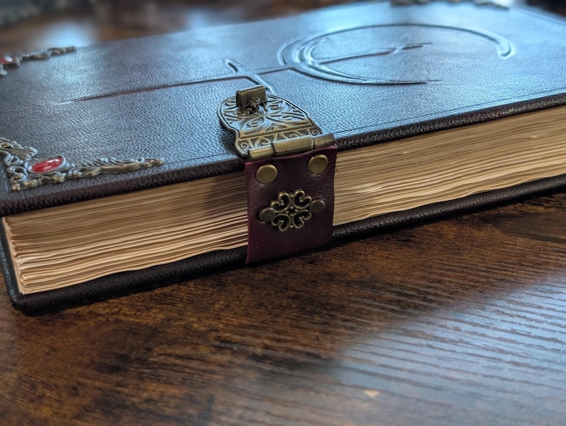 Puede incluir: Un diario de cuero marr&oacute;n oscuro con un cierre y adornos de esquina de metal decorativos. La cubierta presenta un dise&ntilde;o en relieve y las p&aacute;ginas est&aacute;n envejecidas. El diario est&aacute; cerrado con una correa de cuero burdeos.