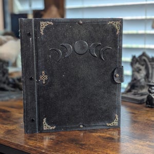 May include: A black leather journal with a triple moon phase design embossed on the cover. The journal features decorative corner accents and a clasp closure. The cover has a textured appearance and a dark, mystical aesthetic.