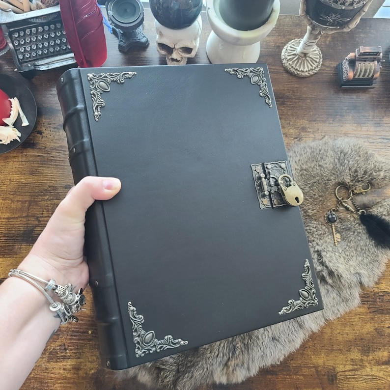 May include: A black journal with a decorative metal clasp and corner accents. The journal has a lock and key, and is held in a person's hand. The journal is on a furry surface.