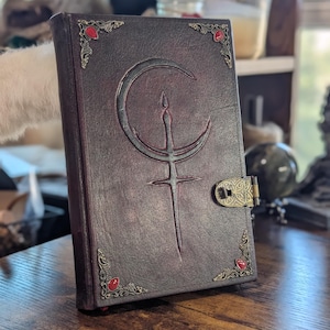 Puede incluir: Un libro encuadernado en cuero marr&oacute;n oscuro con un dise&ntilde;o de luna creciente y espada en relieve en la portada. El libro tiene adornos en las esquinas con gemas rojas y un cierre decorativo. El libro est&aacute; sobre una superficie de madera.