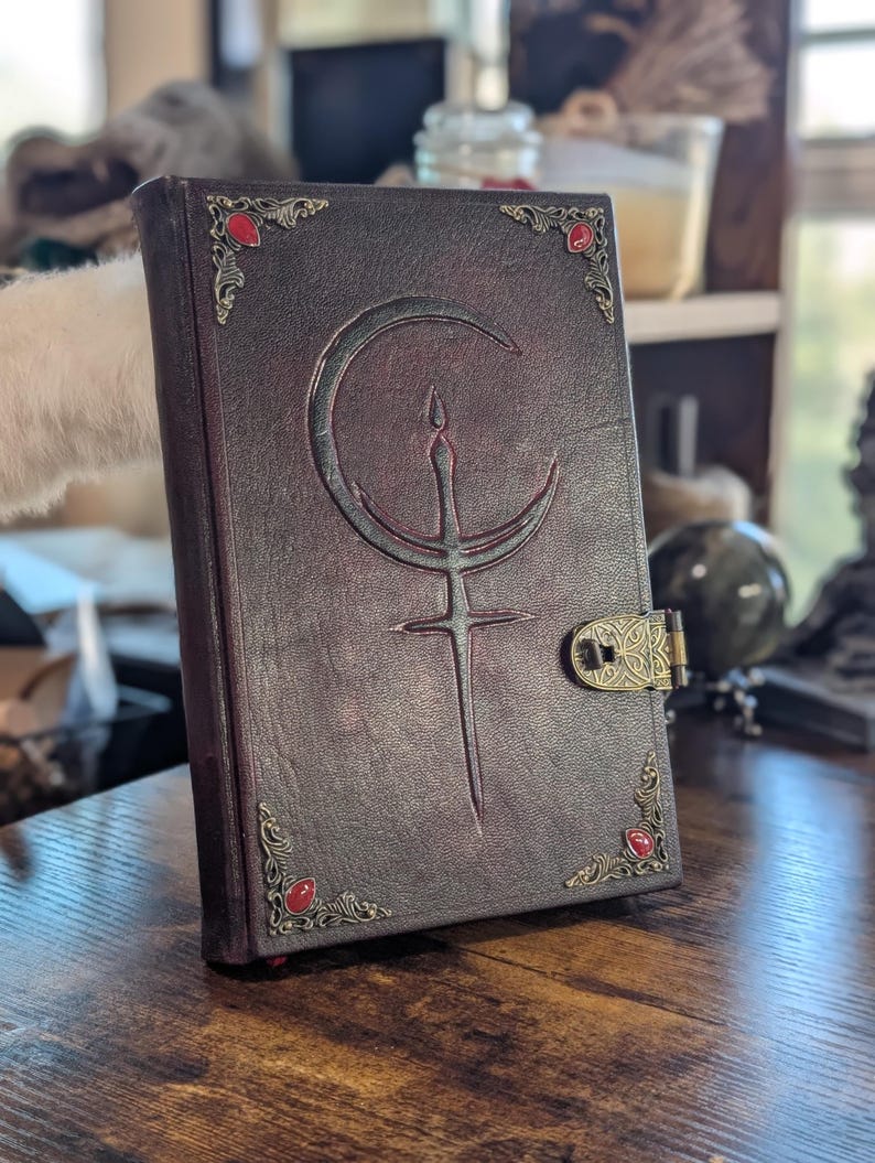 Puede incluir: Un libro encuadernado en cuero marr&oacute;n oscuro con un dise&ntilde;o de luna creciente y espada en relieve en la portada. El libro tiene adornos en las esquinas con gemas rojas y un cierre decorativo. El libro est&aacute; sobre una superficie de madera.
