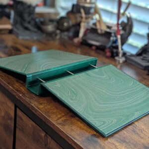 May include: A green marbled desk organizer with a hinged top and metal supports. The organizer is open, revealing the interior. The desk organizer sits on a wooden desk.
