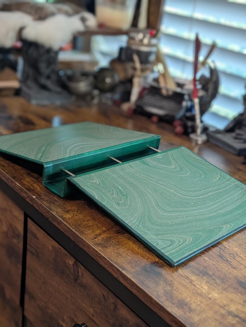 May include: A green marbled desk organizer with a hinged top and metal supports. The organizer is open, revealing the interior. The desk organizer sits on a wooden desk.
