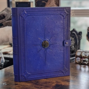 Könnte beinhalten: Ein blaues, ledergebundenes Notizbuch mit Sternenmuster und goldenem Verschluss. Der Einband weist geprägte Details und kleine Sternakzente auf. Das Notizbuch ist geschlossen und scheint neu zu sein.