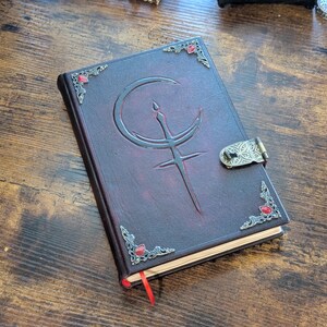 Puede incluir: Un diario de cuero burdeos con un dise&ntilde;o en relieve de luna creciente y espada. El diario presenta adornos en las esquinas con gemas rojas y un cierre decorativo. Se ve una cinta roja como marcap&aacute;ginas.