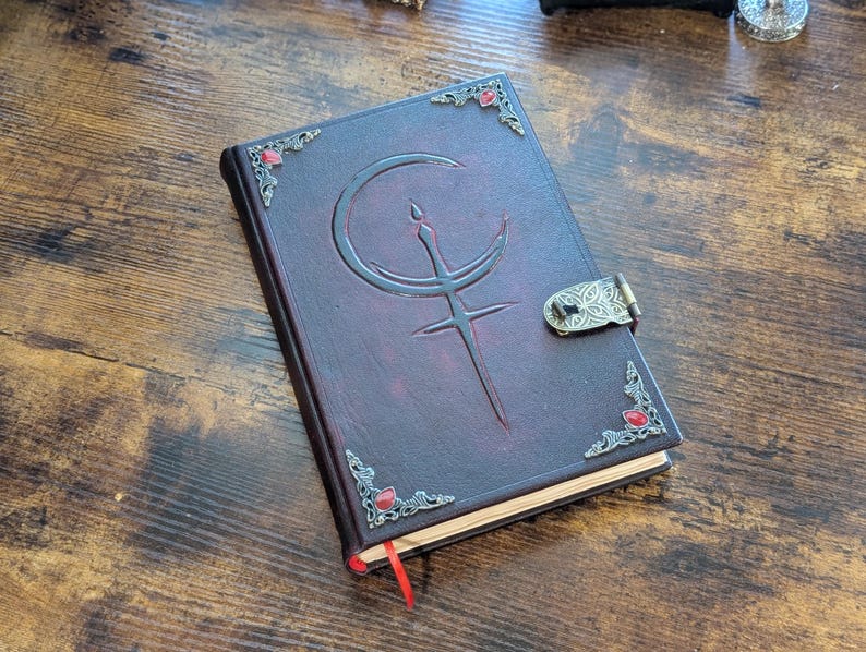 Puede incluir: Un diario de cuero burdeos con un dise&ntilde;o en relieve de luna creciente y espada. El diario presenta adornos en las esquinas con gemas rojas y un cierre decorativo. Se ve una cinta roja como marcap&aacute;ginas.