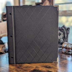 May include: A black photo album with a diamond pattern on the cover. The album has metal corner protectors and a spine with three rivets. The album is sitting on a wooden surface.