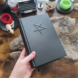 May include: Black leather-bound book with a raised pentagram design. The book has a black ribbon bookmark. The cover is smooth and the edges are squared. The book is held by a person's hand.