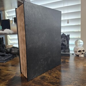K&ouml;nnte beinhalten: Ein gro&szlig;es, schwarzes, ledergebundenes Buch mit einem abgenutzten, alten Aussehen. Das Buch ist geschlossen und steht aufrecht auf einer Holzoberfl&auml;che.