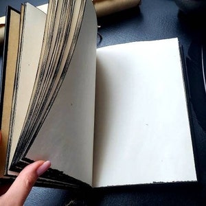 May include: An open, antique-style journal with aged paper pages. The pages are cream-colored with black edges, and the cover is dark. A hand is turning the pages. A quill pen and decorative items are on the dark surface.