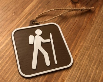 May include: A brown square-shaped ornament with a white hiker silhouette and a walking stick. The ornament has a string attached to it.