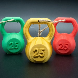 May include: Three miniature kettlebells, one green, one yellow, and one red, are tied together with twine. Each kettlebell has the number "25" printed on it.
