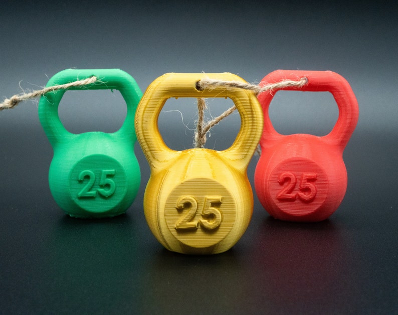 May include: Three miniature kettlebells, one green, one yellow, and one red, are tied together with twine. Each kettlebell has the number "25" printed on it.