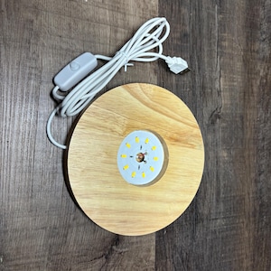 May include: A round, light-colored wooden LED display base with a white USB cable and on/off switch. The base has a circular array of small, yellow LED lights in the center. The USB cable is white.