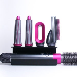 May include: A set of hair styling tools with a pink and black color scheme. The set includes a hairdryer, a round brush attachment, a smoothing brush attachment, and two curling attachments. The tools are arranged on a black stand.