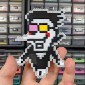 May include: A pixel art character figure crafted from small plastic beads. The figure features a black suit, white shirt, and a prominent nose. It has a yellow square lens over one eye, a pink square lens over the other, and a red cheek mark.