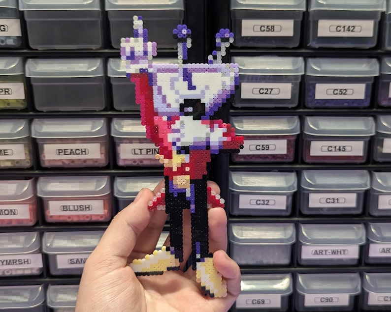 May include: A pixel art figure crafted from small plastic beads, depicting a character with a white cup-shaped head, a red jacket, black pants, and yellow shoes. The figure is held upright, showcasing a vibrant color palette and a grid-like texture.