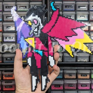 May include: A pixel art figure made of fused plastic beads depicting a character with a white face, black hair, and a large pink and yellow wing. The figure wears a dark suit with a blue tie and has a white arm extended upward.