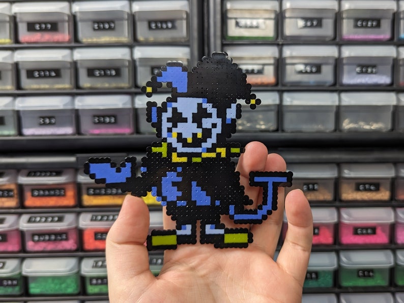 May include: A pixel art figure crafted from small plastic beads, depicting a jester character with a light blue face, a black and blue hat, a black suit, and lime green accents on the collar and shoes, featuring a wide, toothy grin.