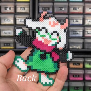 May include: A pixel art character figure crafted from small, fused plastic beads. The figure features a white head with pink horns, a dark pink scarf, a green tunic, and black limbs. The word Back is printed in white text on the lower portion.