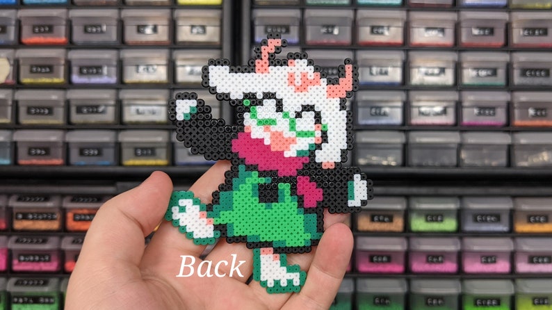 May include: A pixel art character figure crafted from small, fused plastic beads. The figure features a white head with pink horns, a dark pink scarf, a green tunic, and black limbs. The word Back is printed in white text on the lower portion.