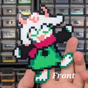 May include: A pixel art figure made of fused plastic beads depicting a character with white fur, green glasses, a pink scarf, a green tunic, and black sleeves. The character is posed with one arm raised and one leg lifted, featuring the text Front.