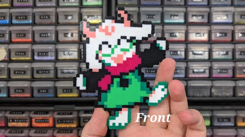 May include: A pixel art figure made of fused plastic beads depicting a character with white fur, green glasses, a pink scarf, a green tunic, and black sleeves. The character is posed with one arm raised and one leg lifted, featuring the text Front.