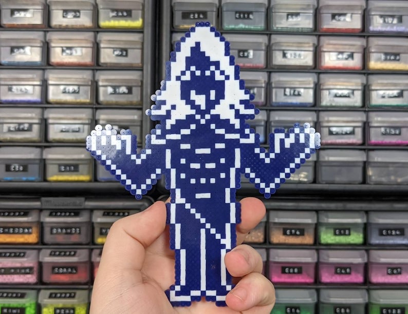 May include: A pixel art figure crafted from dark blue and white plastic fuse beads. The design depicts a stylized humanoid character with a pointed head, raised arms, and a long robe-like garment, constructed in a grid-like pattern of small beads.
