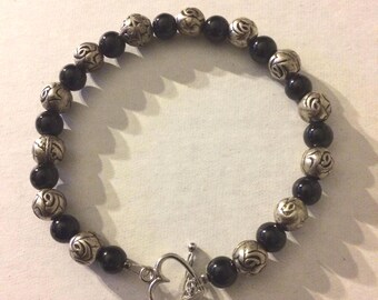 May include: A beaded bracelet featuring alternating black and silver beads. The silver beads have a floral design. The bracelet is secured with a silver heart-shaped clasp. The bracelet is approximately 7 inches in circumference.