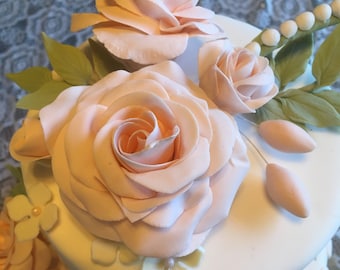 May include: A three-tiered wedding cake decorated with delicate pink sugar roses and green leaves. The cake is white with a light pink ribbon around the bottom tier.